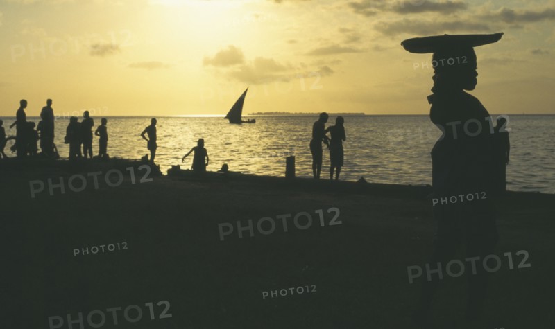 TANZANIA , Zanzibar, Zanzibar, People on the waterfront silhouetted against yellow sunset with dhow at sea in the distance