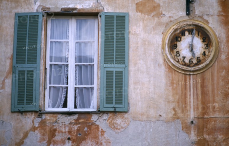 FRANCE, Provence Cotes d’Azur , Alpes-Maritimes, "Sospel.  Architectural detail, clock and window with open green shutters set into outside wall of building with faded, crumbling terracotta plaster."