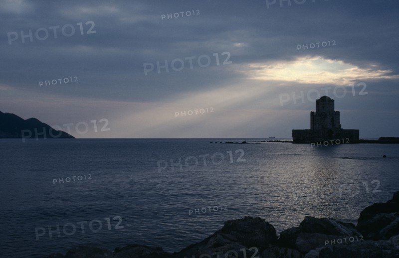 GREECE, Peloponnese, Methoni, View across the sea towards fortress ruins lit by evening sun rays coming through a break in the clouds.