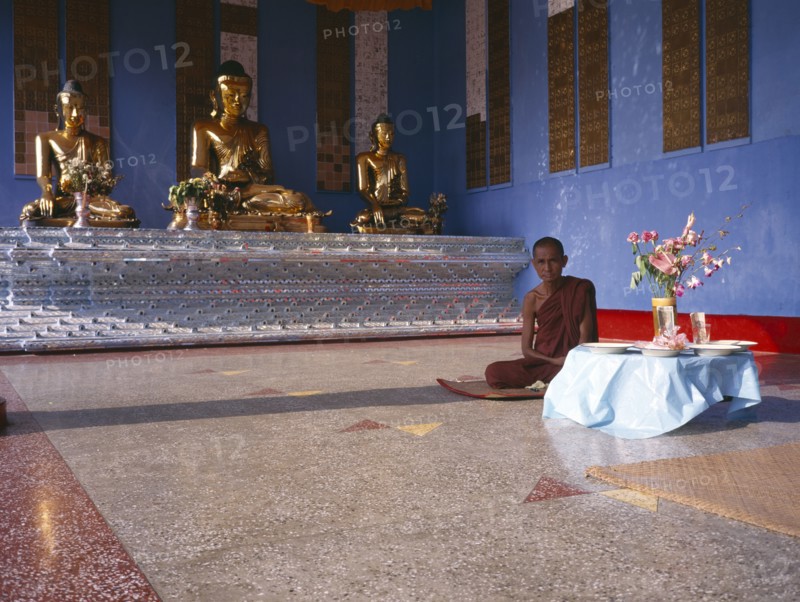 MYANMAR, Yangon, "Shwedagon Pagoda interior.  Monk sitting beside a raised altar with three seated, golden Buddha figures.   "