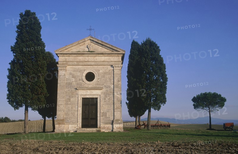 ITALY, Tuscany, San Querico, "Small stone chapel, facade with cypress trees growing beside it in an arable landscape."