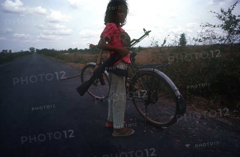 CAMBODIA, Kompong Thom, Ten year old girl standing on the Route 6 roadway beside her bicycle holding an AK47.