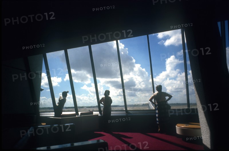 USA, Florida, Miami, Miami Airport. Three figures looking out of window towards planes framed by interior walls in heavy shadow.
