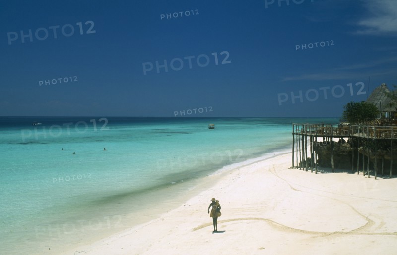 TANZANIA, Zanzibar Island, "Nungwi.  Single figure walking along sandy beach beside aquamarine water.  Beach huts, tables and chairs on raised wooden platform on righthand side."