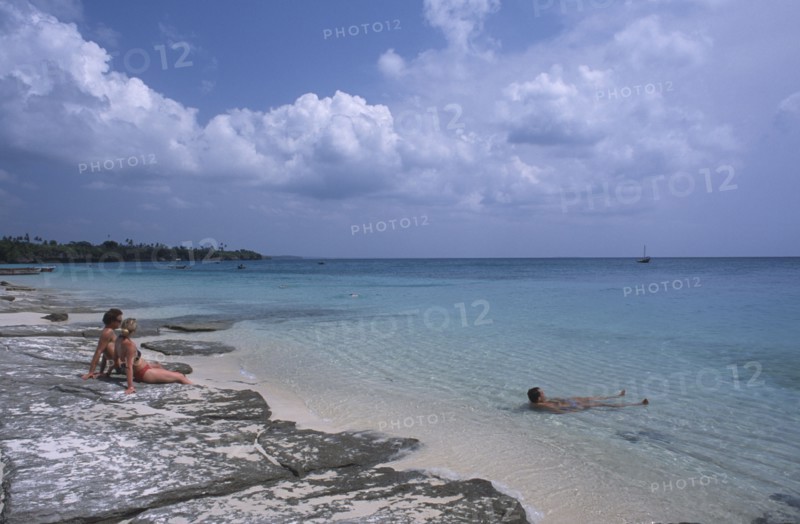 TANZANIA, Zanzibar Island, Mangapwani, Young couple sitting on flat rocks on the shoreline looking out over aquamarine sea with another man lying in the shallow water.
