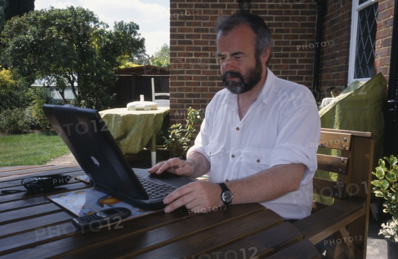COMPUTERS, Laptop, Man sitting at a table outdoors using a laptop linked to NFT via a mobile phone.
