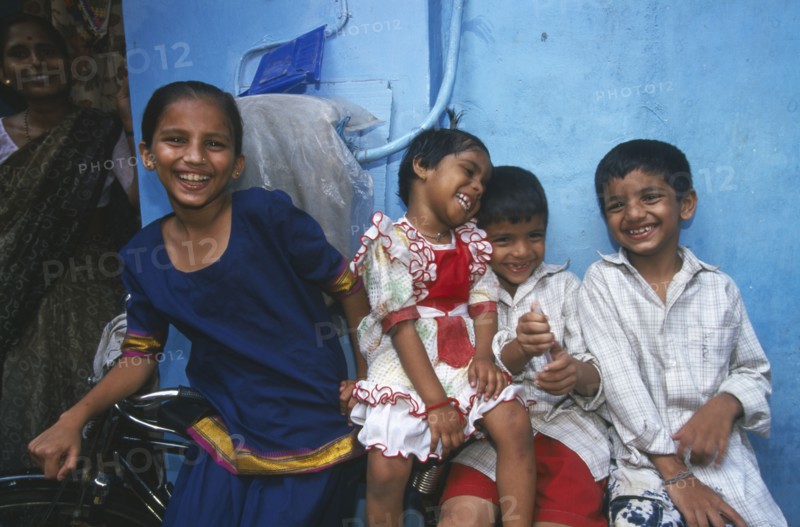 INDIA, Maharashtra, Mumbai, Four smiling children leaning against bicycle with blue wall behind.