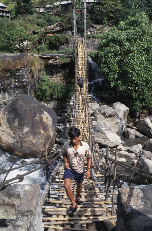 NEPAL, Annapurna Region, Marsyangdi River, Near Besisahar.  Young man crossing wood and rope foot bridge over rocky river gorge.  With person carrying backpack in the opposite direction at far end.