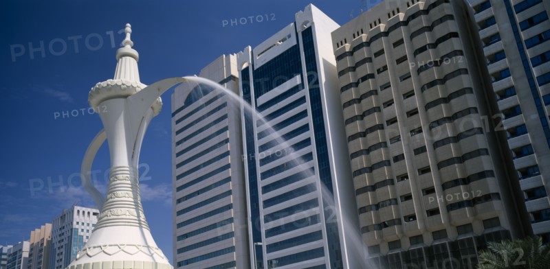 UAE, Abu Dhabi, Al-Ittihad Square. Fountain of water falling from the spout of a sculpted Arabian coffeepot or dallah with high rise buildings behind.