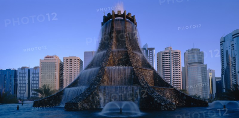 UAE, Abu Dhabi, The Volcano Fountain. Large tiered fountain with water cascading from circular top and high rise modern buildings behind.