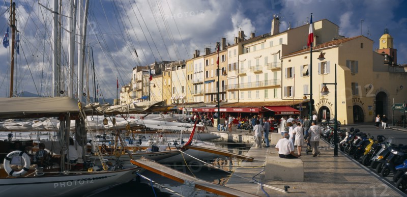 FRANCE, Provence-Cote d’Azur, St Tropez, "View along busy waterfront  with boats moored at the harbourside, open-fronted restaurants and the local tourist office.  Var region."