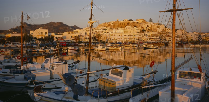 GREECE, Cyclades Islands, Naxos, Hora.  View over moored boats and harbour towards town reflected in the water in evening light.