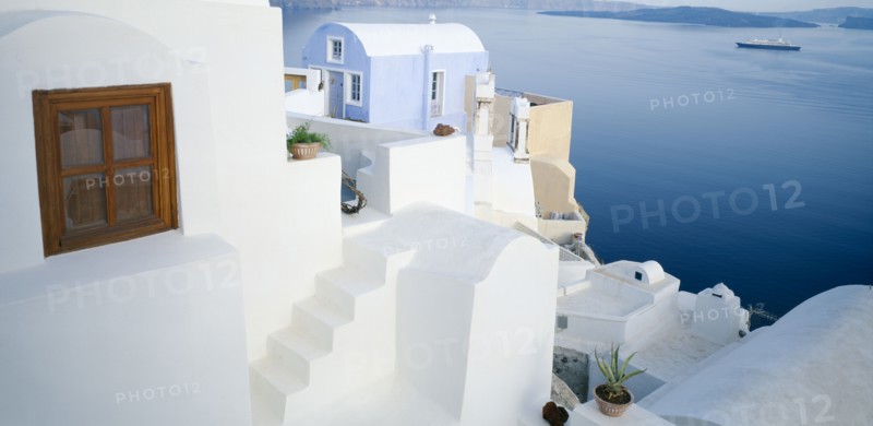 GREECE, Cyclades Islands, Santorini, Oia.  White painted terrace and roof tops overlooking sea with distant ferry and coastline behind.