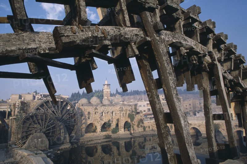 SYRIA, Central, Hama, Wooden norias or waterwheels on the Orontes river.  Part view of wheel section in the foreground framing the Al-Nuri Mosque dating from 1172 and built of limestone and basalt.