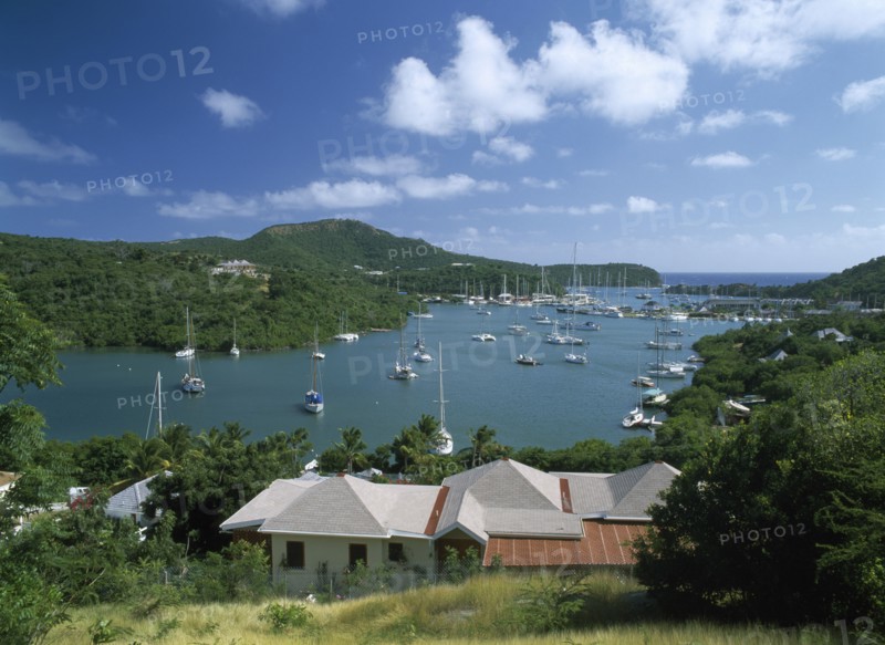 WEST INDIES, Antigua, English Harbour, Ordnance Bay.  View over rooftops towards bay with moored yachts and surrounding tree covered coastline.