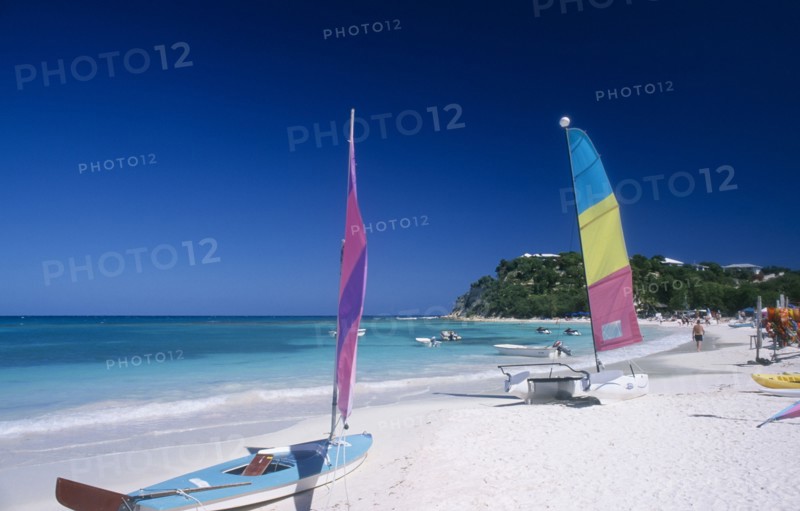 WEST INDIES, Antigua, Long Bay, "Semi circular bay beside quiet beach, boats with colourful sails pulled up onto the sand and motor boats and jet skis moored in the water.  Tree covered headland behind. "