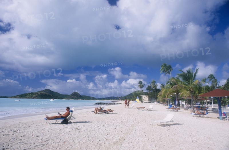WEST INDIES, Antigua, Jolly Beach, View along sandy beach lined with palm trees and sun umbrellas towards hilly coastline.  People sunbathing on loungers in the foreground.