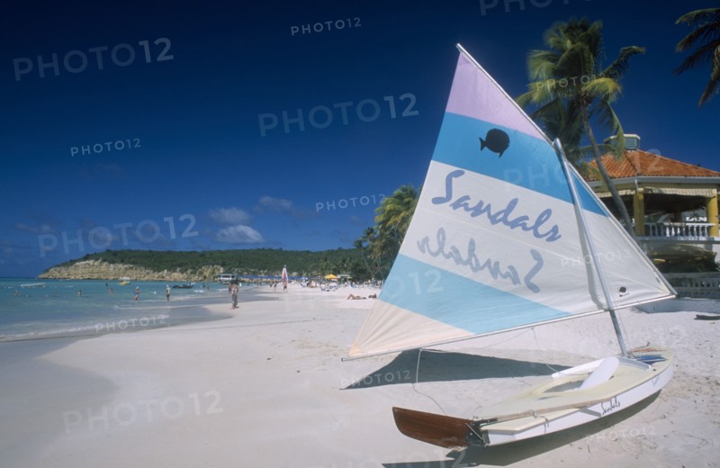 WEST INDIES, Antigua, Dickenson Bay, View along sandy beach lined with palms with boat in the foreground advertising Sandals hotel on its hull and sail.  Building rooftop part seen behind.