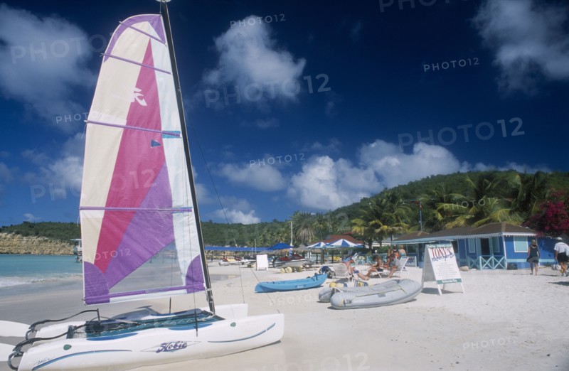 WEST INDIES, Antigua, Dickenson Bay, "Sandy beach with people on deck chairs beside blue painted building, A-board advertising Tonys watersports.  Hobiecat with bright sail in front."