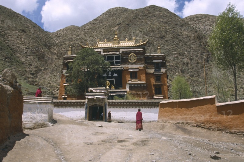 CHINA, Gansu, Xiahe, "Labrang Monastery, elaborately designed building with a golden roof, partially obscured by a tree, and monks walking in the foreground."