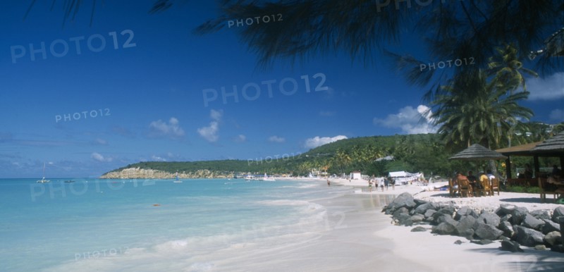 WEST INDIES, Antigua, Dickenson Bay, View along beach towards tree covered headland with people walking along shoreline and sitting under thatched sun umbrellas nearby.