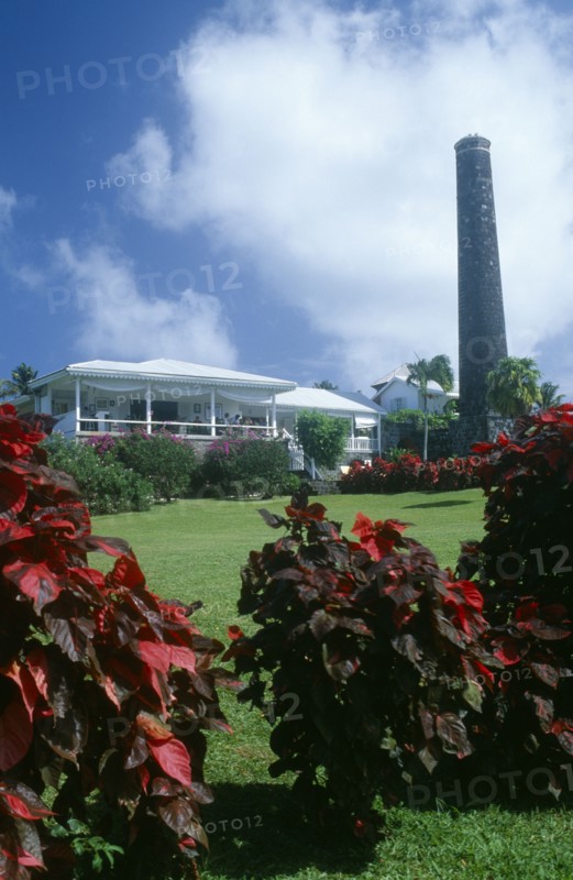 WEST INDIES, St Kitts, Rawlins Plantation Inn, Exterior with people on white painted balcony and old sugar factory tower and wall on the right.  Red and purple variegated plants in the foreground.