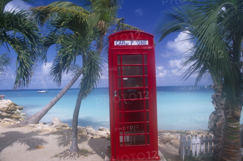 WEST INDIES, Antigua, Dickenson Bay, Red telephone box on sand surrounded by palm trees with sea and distant boat behind.