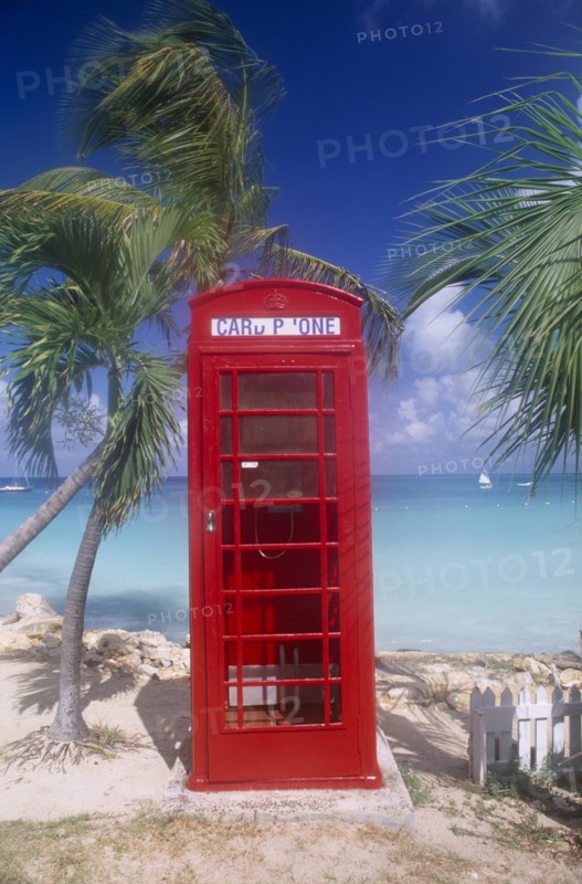 WEST INDIES, Antigua, Dickenson Bay, Red telephone box on sand surrounded by palm trees with sea behind.