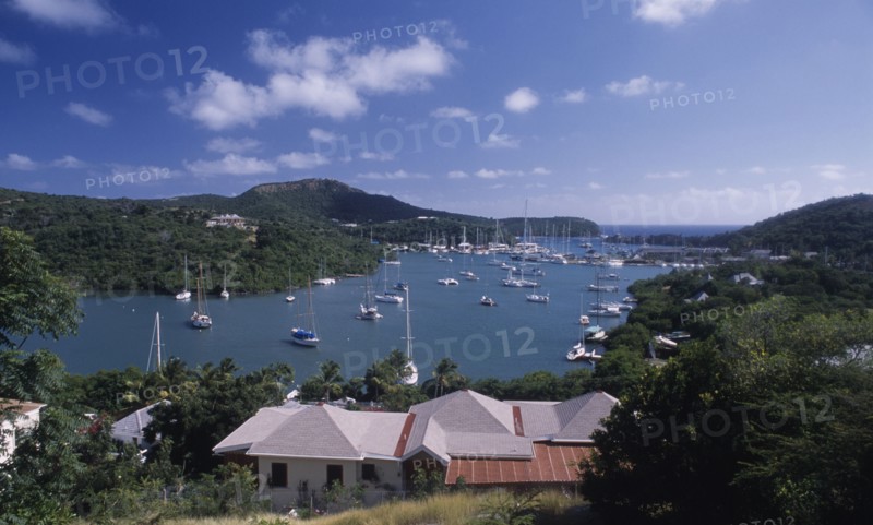 WEST INDIES, Antigua, Ordnance Bay, View over house rooftops towards bay with moored yachts and surrounding coastline.