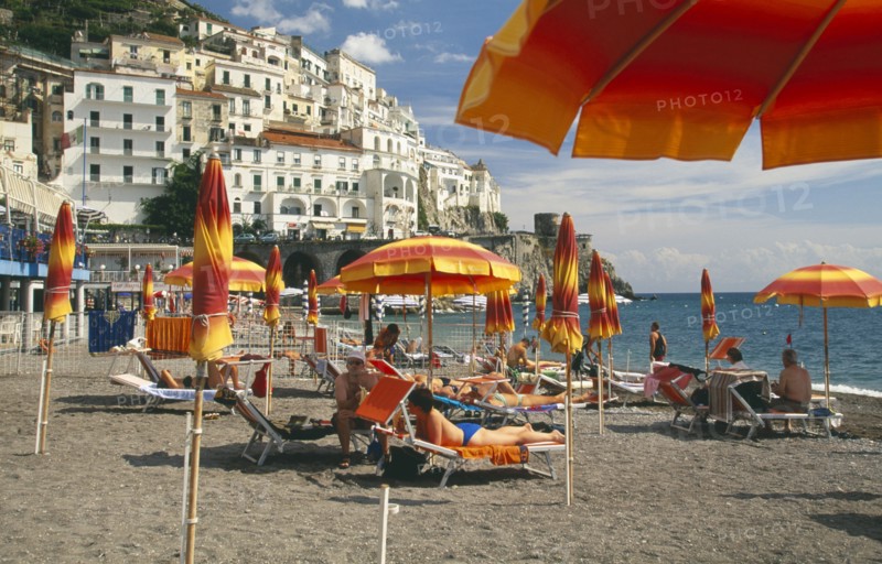 ITALY, Campania, Amalfi Coast, Amalfi.  Beach with sunbathers on loungers under yellow and orange striped beach umbrellas.  Town buildings on the hillside behind.