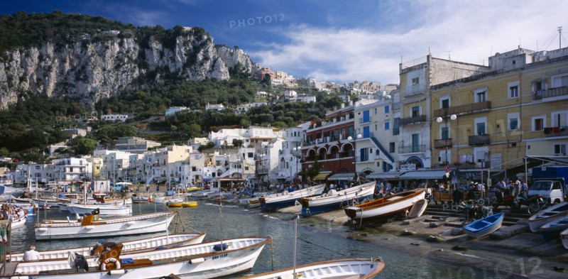 ITALY, Campania, Capri, Marina Grande.  View over moored boats in harbour lined with colourful houses and busy cafes and restaurants.  Rocky hillside behind.