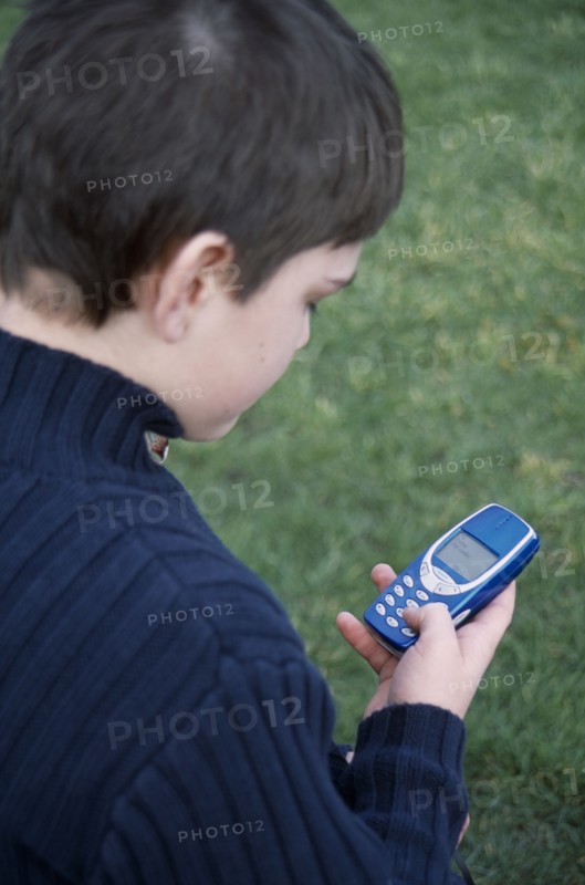 COMMUNICATIONS, Telephones, Mobile, View over the shoulder of a boy holding a blue mobile phone with his thumb on the dialing pad.