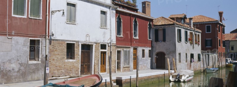 ITALY, Veneto, Venice, Murano Island.  View along canalside houses with boats moored to posts in front.