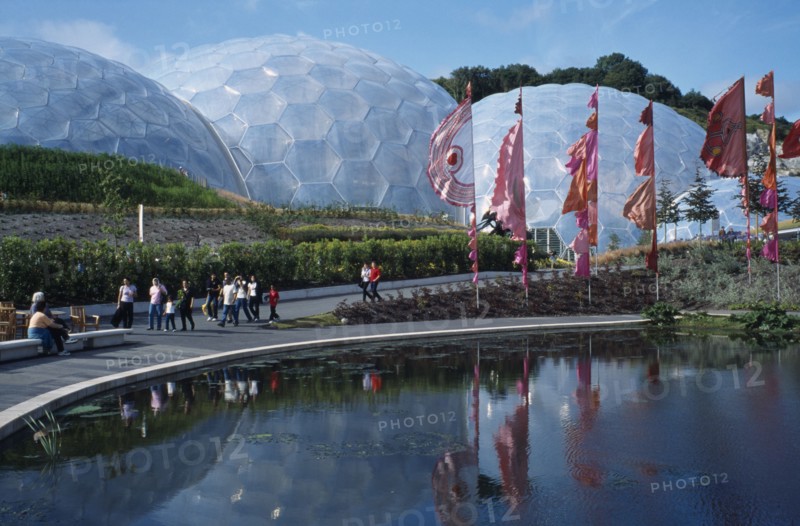 ENGLAND, Cornwall, St. Austell, Eden Project.  General view over the Humid tropics Biome exterior with line of coloured flags and visitors walking beside lake.