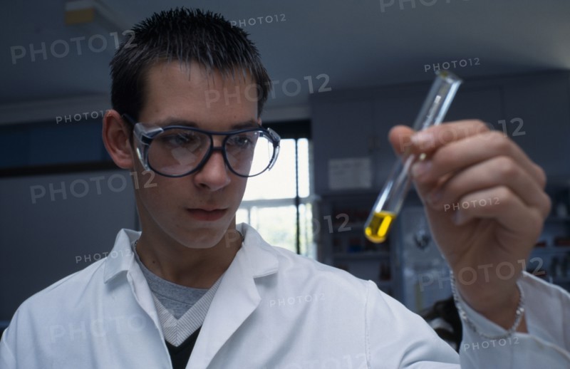 EDUCATION, Science Class, Male student wearing protective glasses and lab coat holding a test tube containing yellow liquid at an angle.