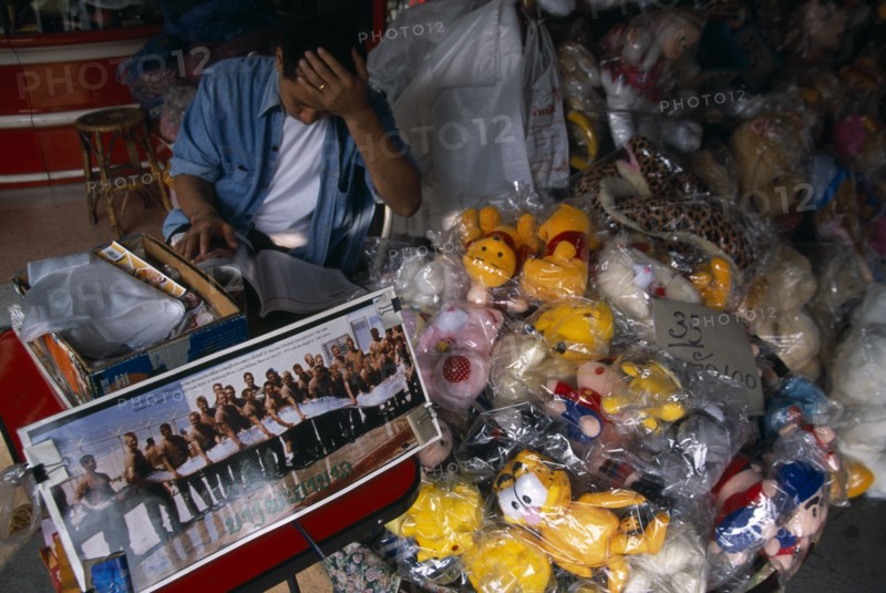 THAILAND, North, Chiang Mai, Chinatown. Shop with plastic wrapped toys for sale and picture commemorating the capture of a huge fish by the American Army in 1973.  Male vendor reading behind.
