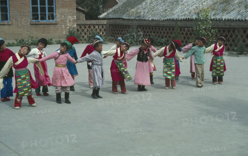 CHINA, Hebei Province, Chengde, Line of children at kindergarten with linked arms and heads inclined sideways wearing traditional dress.
