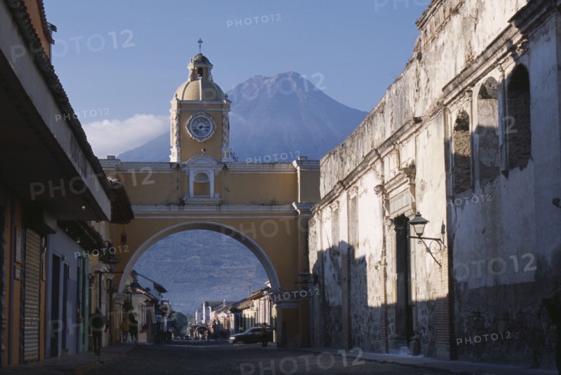 GUATEMALA, Antigua, "Arco de Santa Catarina. Yellow painted archway with clock tower and people walking underneath, with Volcan Agua beyond."