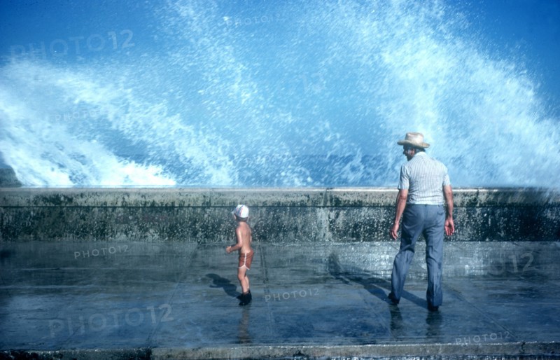 CUBA, Havana, Malecon, Old man and small boy standing on pavement with waves crashing over the sea wall