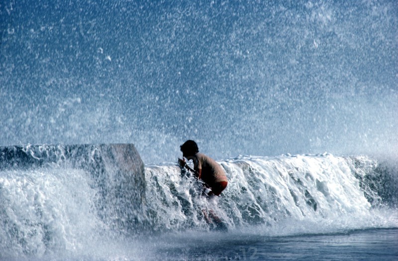 CUBA, Havana, Malecon, Young boy standing on pavement with waves crashing over the sea wall