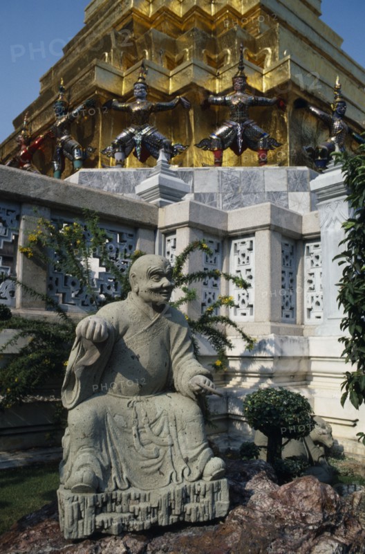 THAILAND, Bangkok, Grand Palace, Aka Wat Phra Kaeo. Stone statue on rockery next to small tree in grounds