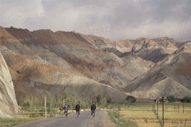 CHINA, Quinghai Province, Near Guide, Row of three cyclists on a road leading toward rocky mountain landscape