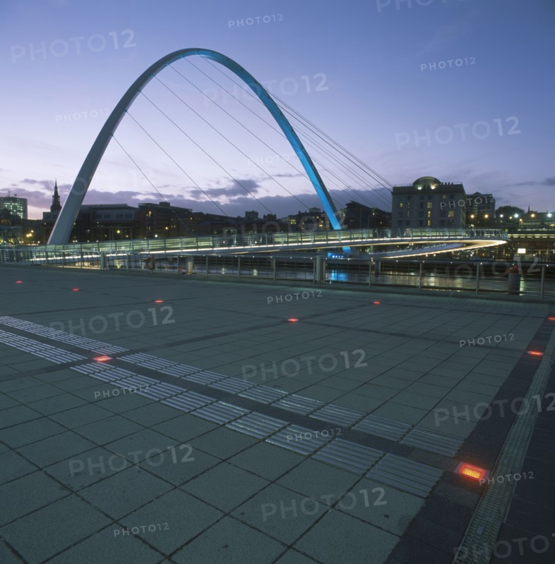 ENGLAND, Tyne and wear, Gateshead, The new Millennium Footbridge illuminated at dusk with city buildings silhouetted against purple evening sky behind.