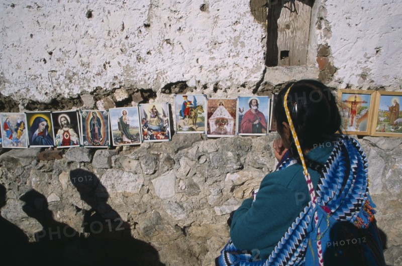 MEXICO, Chiapas, San Cristobal de Las Casas, Woman in front of coloured pictures of the lives of the Saints and Christ displayed along stone wall.
