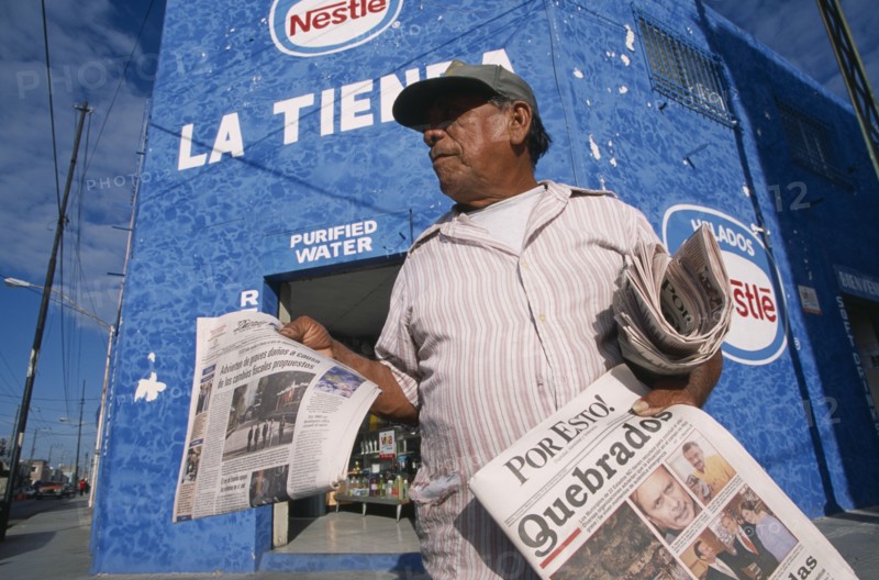 MEXICO, Yucatan, Merida, "Male newspaper vendor, threequarter view seen from below looking up.  Advertising for Nestle and purified water on blue painted wall of building behind. "