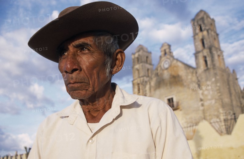 MEXICO, Yucatan, Hoctun, "Elderly man in hat, head and shoulders portrait with church building part seen in background."