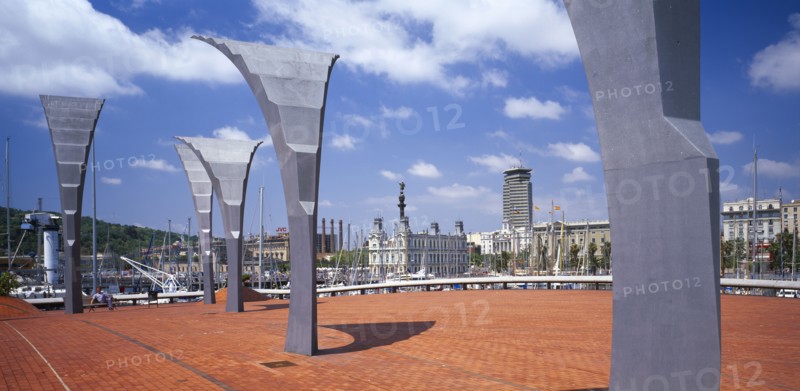 SPAIN, Catalonia, Barcelona, Port Vell.  Modern sculpture on paved area with masts of moored yachts seen behind and statue of Christopher Columbus seen above city buildings beyond.