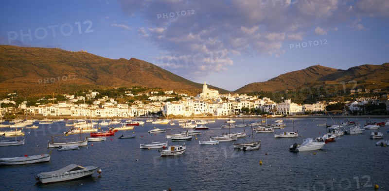 SPAIN, Catalonia, Cadaques, View over moored boats towards white painted town at foot of steep hillside.