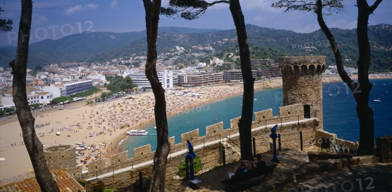 SPAIN, Catalonia, Tossa de Mar, View over busy sandy beach and town from fortified walls of Villa Vella with line of trees in the foreground.