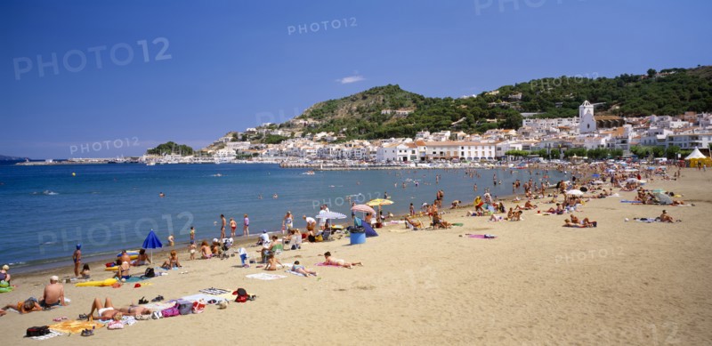 SPAIN, Catalonia, El Port de La Selva, "Curved, sandy beach with people sunbathing and in the water with white painted town at foot of tree covered hillside beyond. "